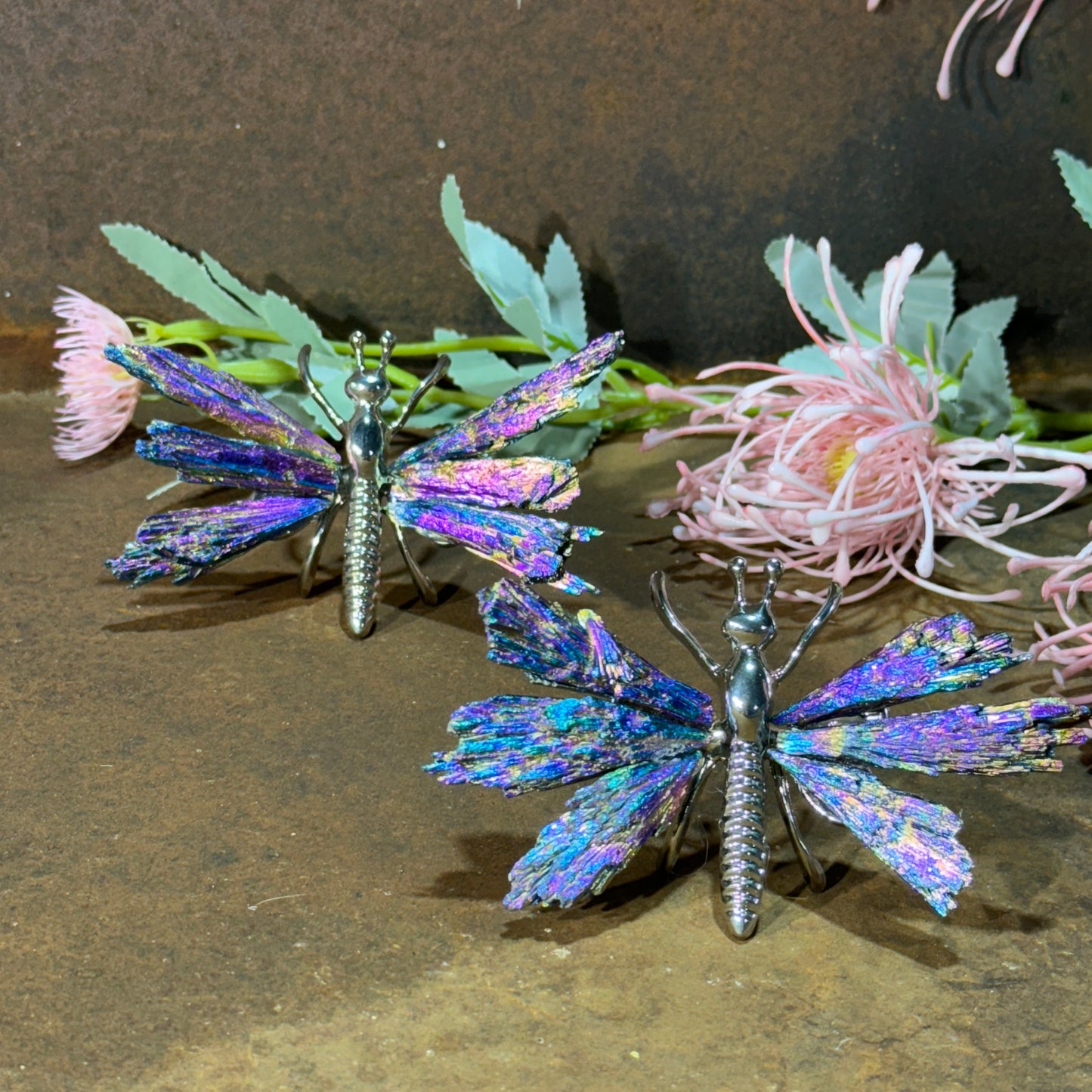 A crystal butterfly with a metallic structure and a multicolored, shimmering aura coating, displayed on a wooden surface with pink floral decorations in the background.