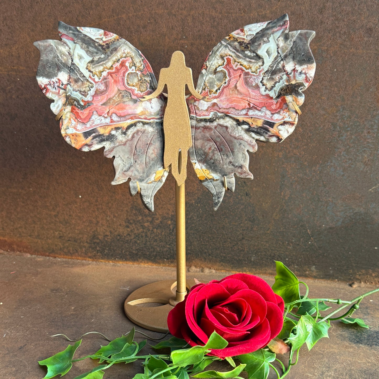 A decorative piece featuring wings made from Mexican Agate on a stand, with a artificial red rose and green leaves in front, set against a rustic background.