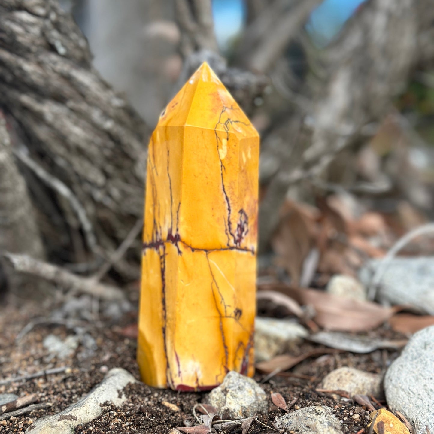 A yellow and red mookaite jasper crystal tower standing upright on a natural ground with trees in the background.