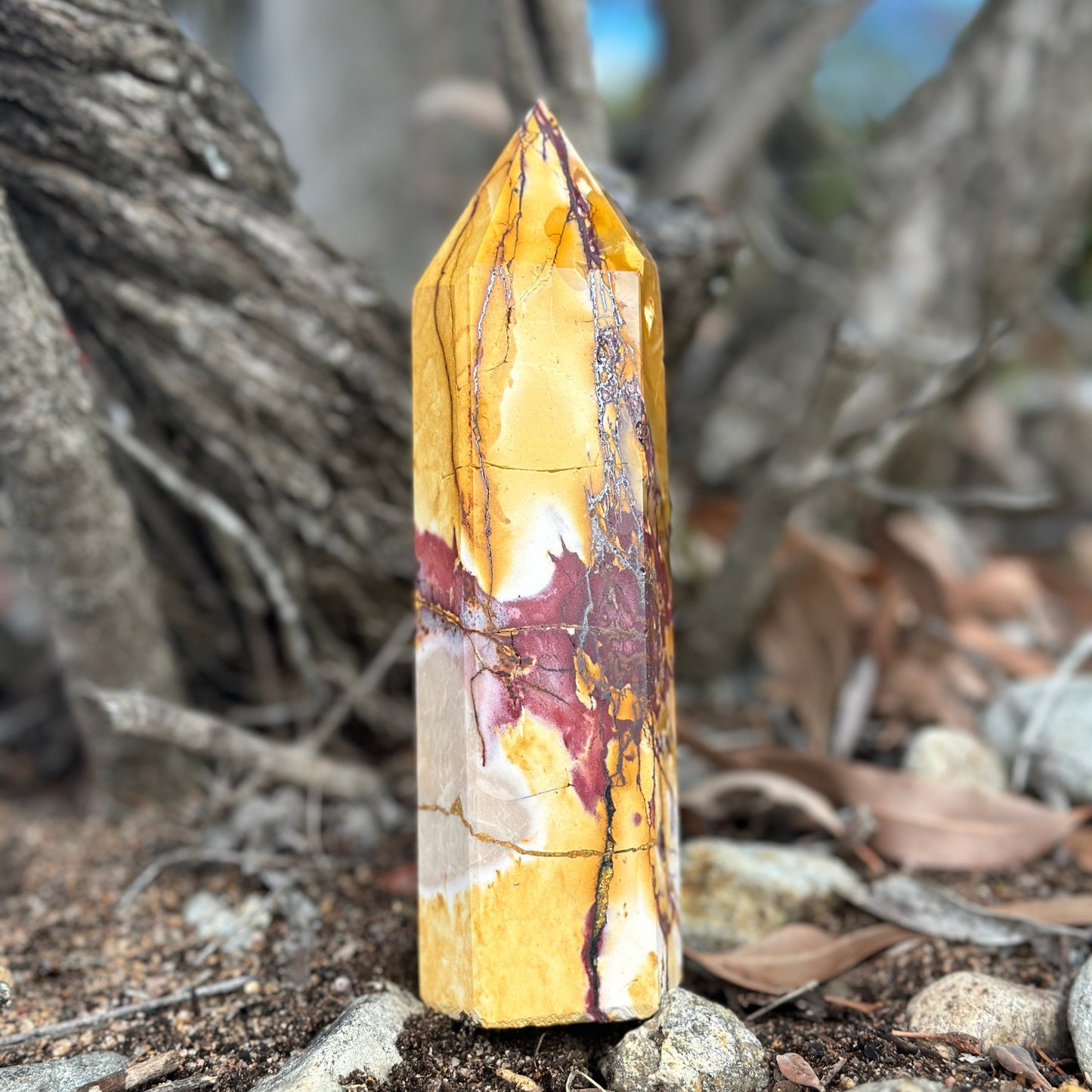 A yellow and red mookaite jasper crystal tower standing upright on a natural ground with trees in the background.