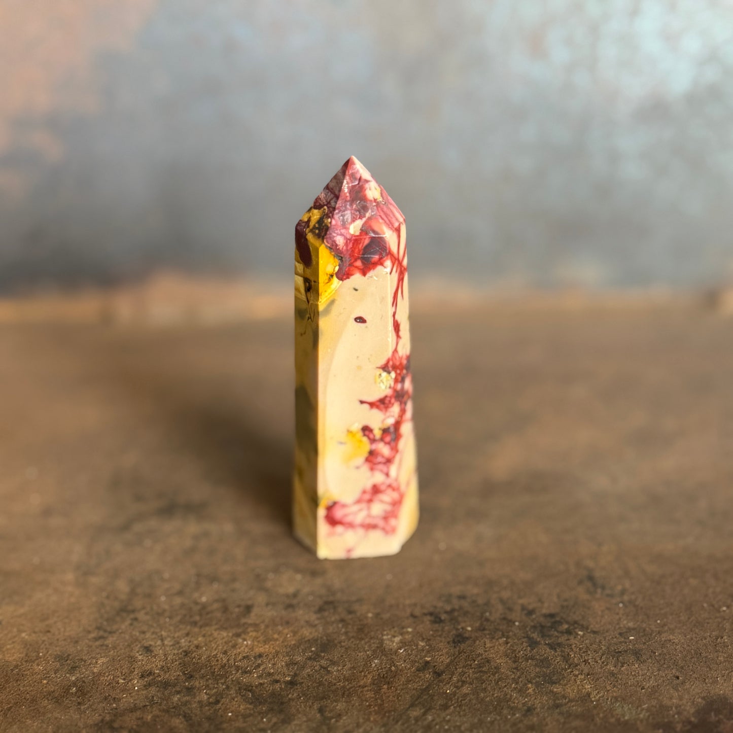 A yellow and red mookaite jasper crystal tower standing upright on a natural ground with trees in the background.