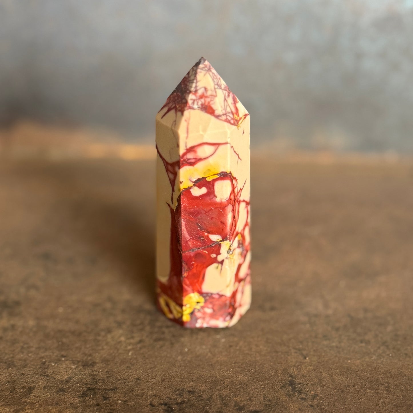A yellow and red mookaite jasper crystal tower standing upright on a natural ground with trees in the background.