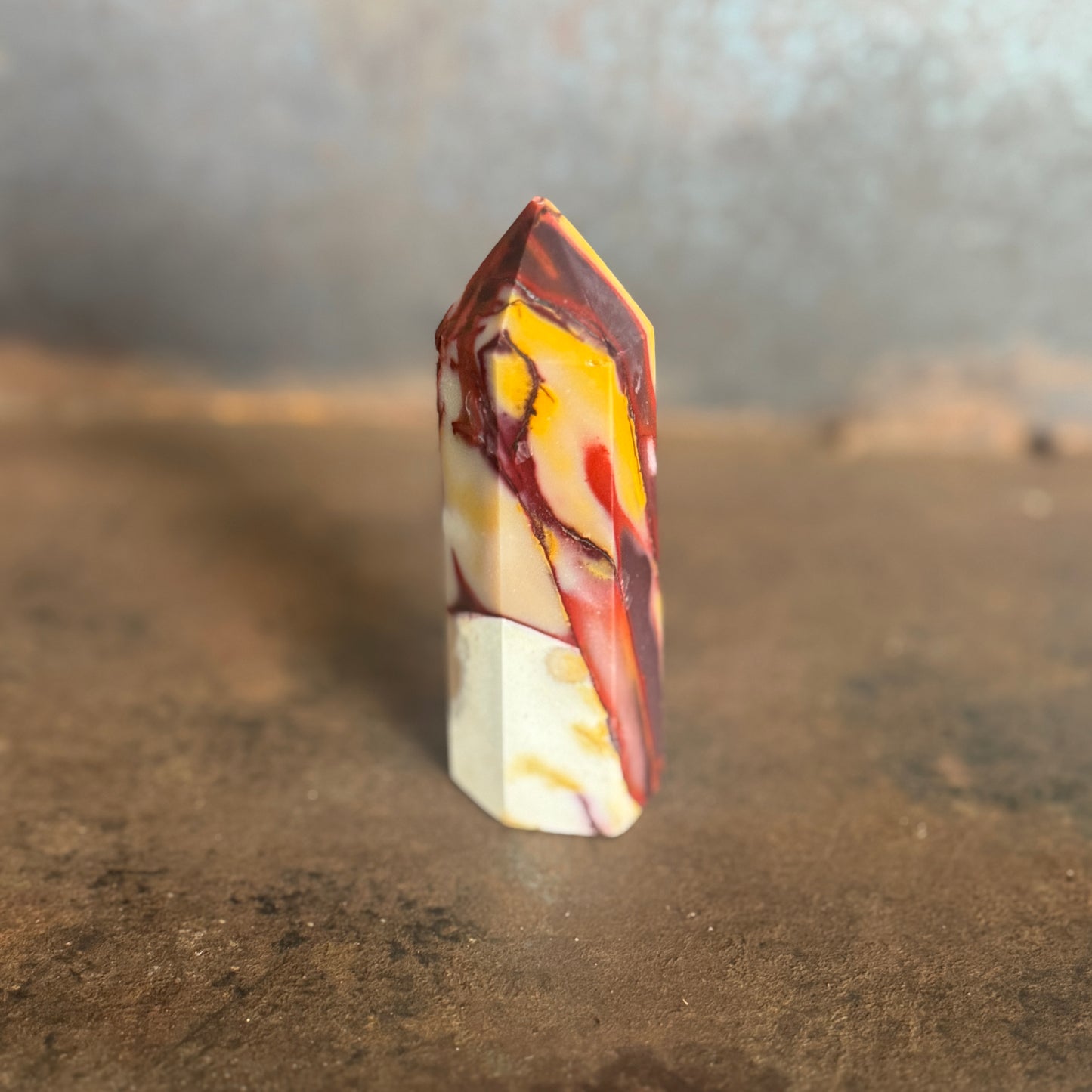 A yellow and red mookaite jasper crystal tower standing upright on a natural ground with trees in the background.