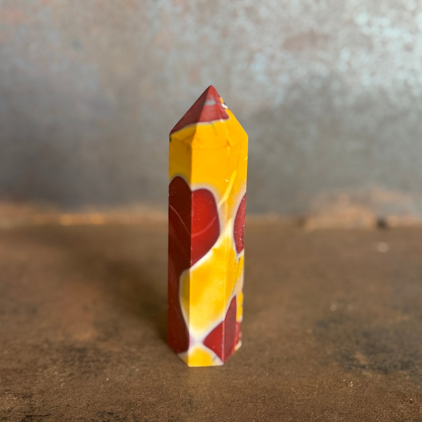 A yellow and red mookaite jasper crystal tower standing upright on a natural ground with trees in the background.