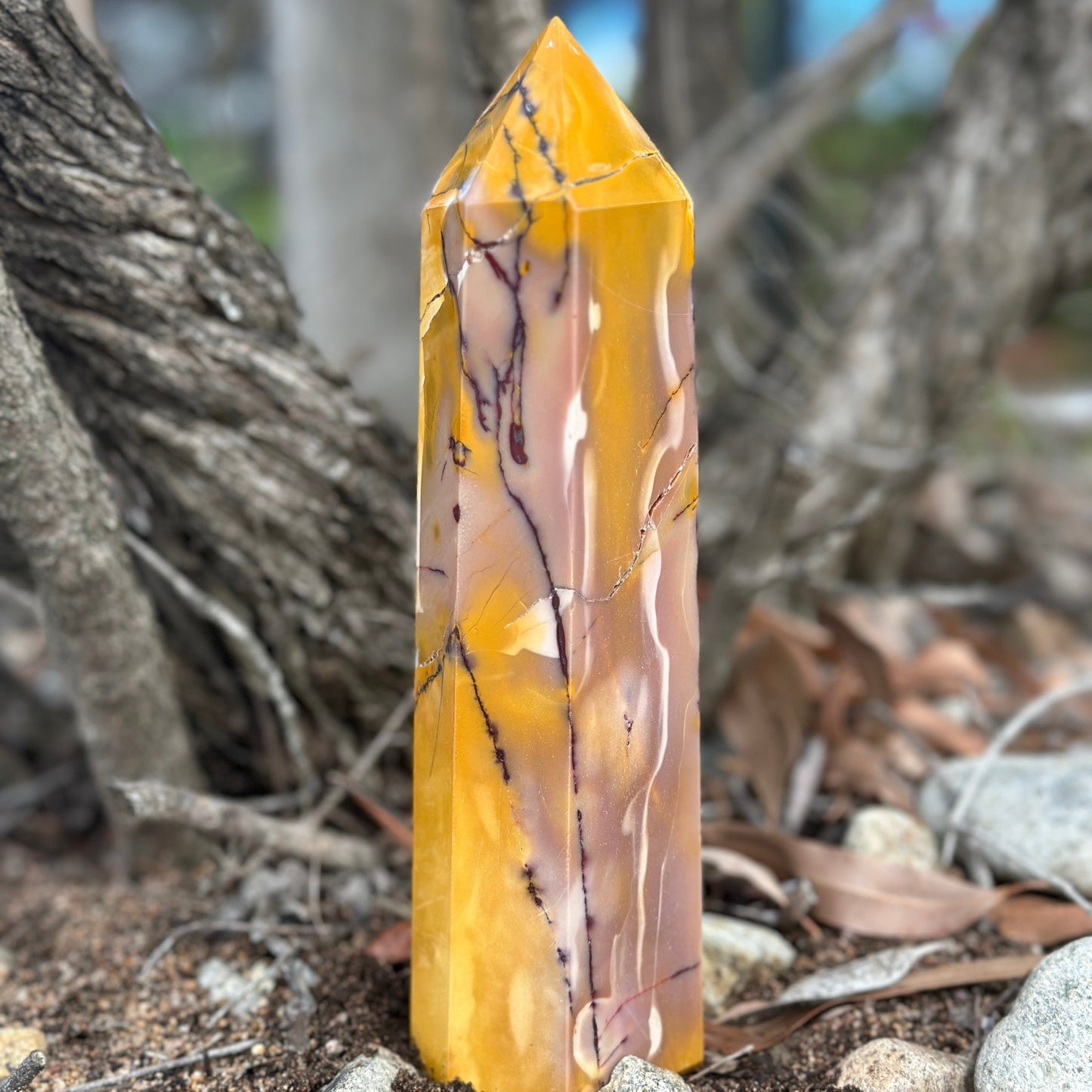 A yellow and red mookaite jasper crystal tower standing upright on a natural ground with trees in the background.