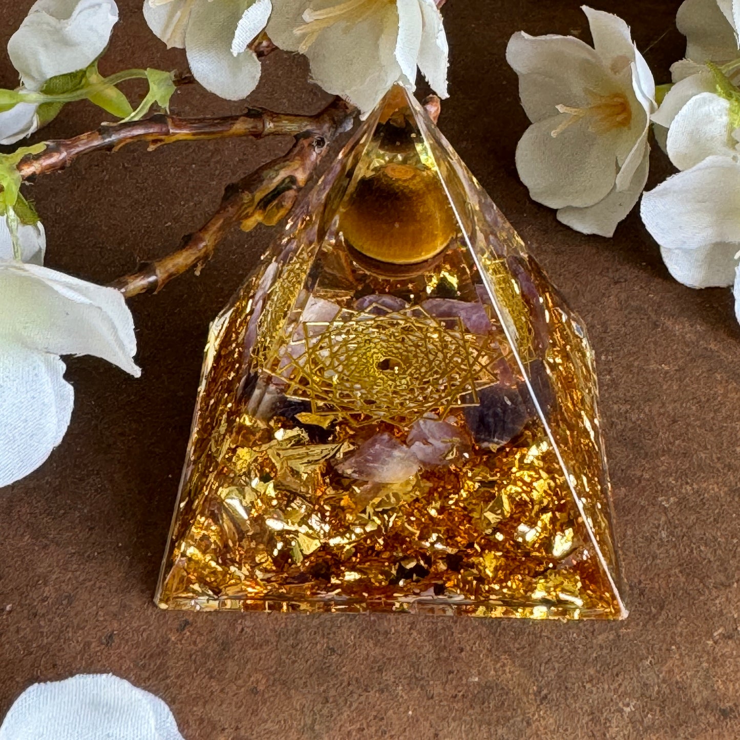 A golden Orgonite Pyramid with Amethyst and Tiger's Eye, adorned with a Flower of Life design, set against a neutral background with white flowers.