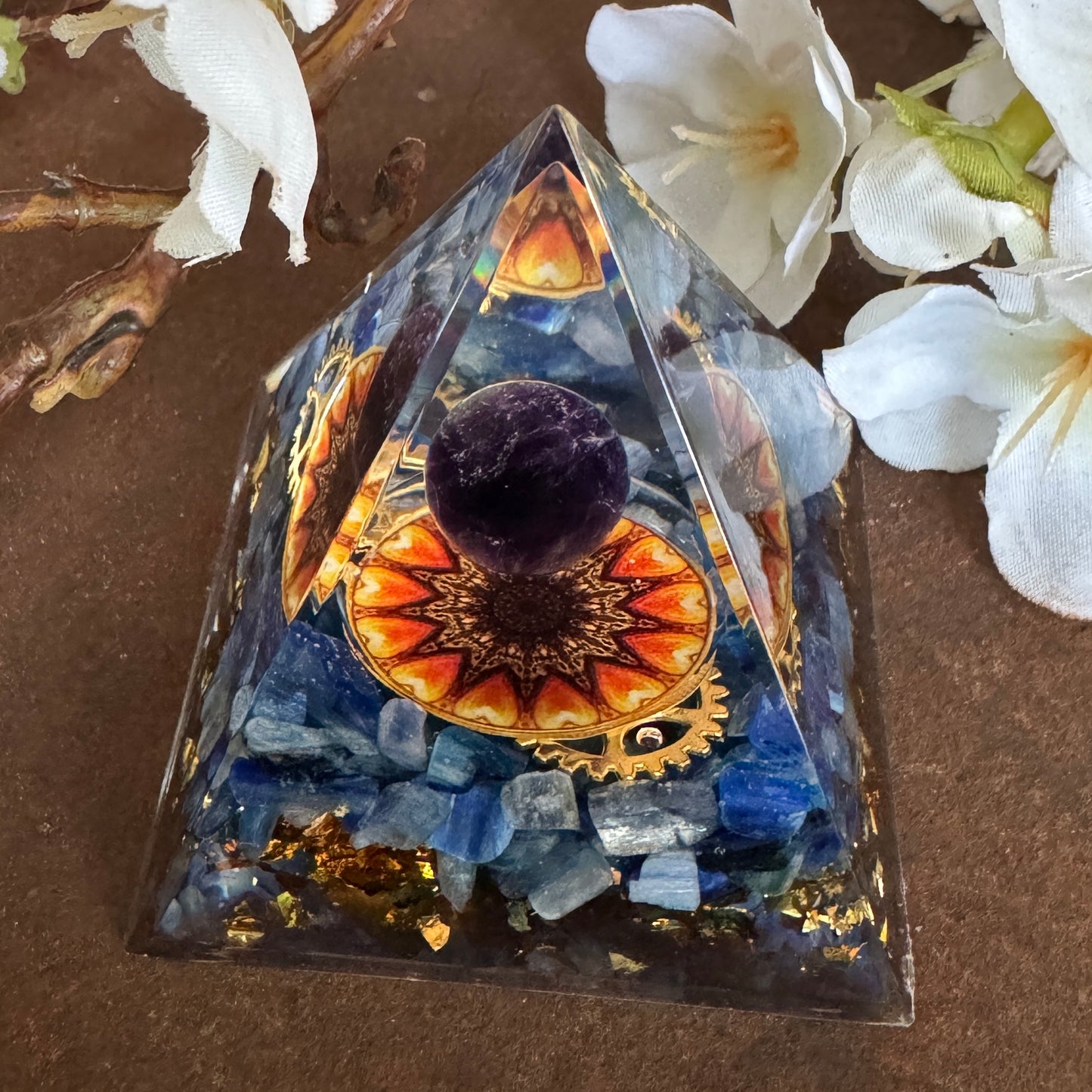 A triangular Orgonite Pyramid with a central Amethyst sphere and Sun feature, surrounded by Kyanite and set against a floral backdrop.