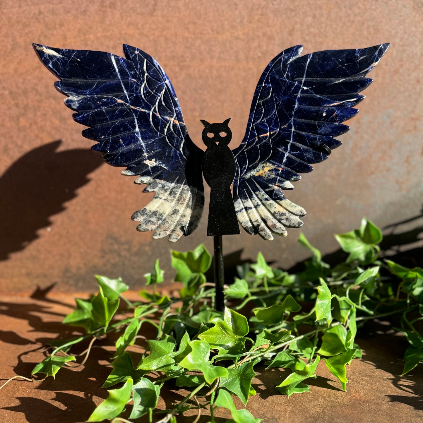 A sodalite stone owl stand with blue wings displayed against a brown background with green leaves in the foreground.