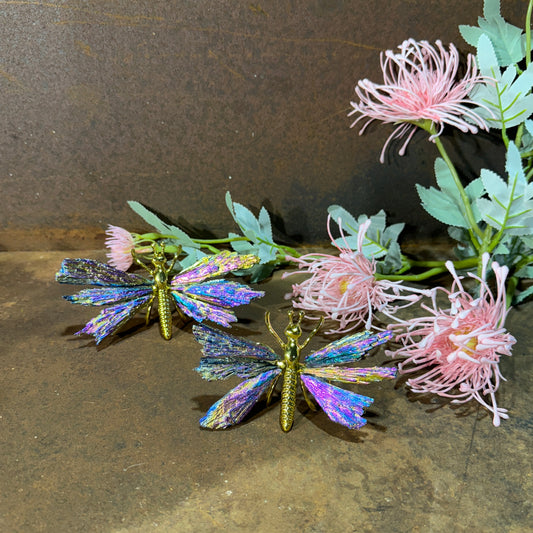 A crystal butterfly with a metallic structure and a multicolored, shimmering aura coating, displayed on a wooden surface with pink floral decorations in the background.