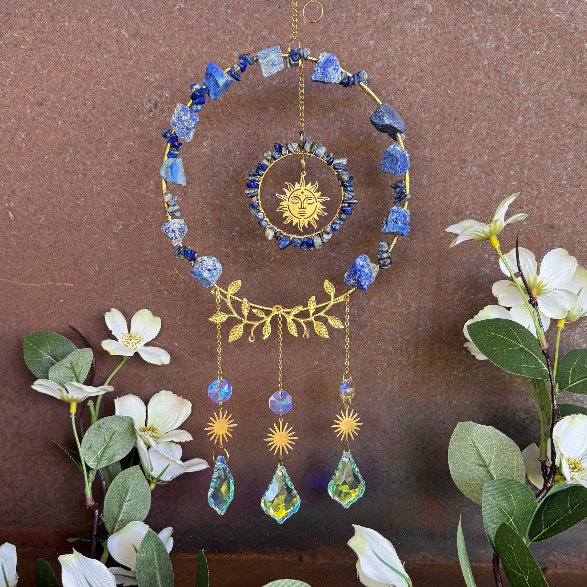 Decorative crystal hanging ornament with blue stones, gold accents, and crystal-like elements on a brown background with flowers.