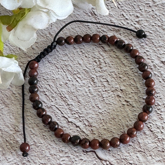 Adjustable bracelet made of Mahogany Obsidian beads, displayed on a wooden-like surface with white flowers in the background.