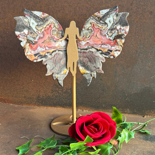 A decorative piece featuring wings made from Mexican Agate on a stand, with a artificial red rose and green leaves in front, set against a rustic background.