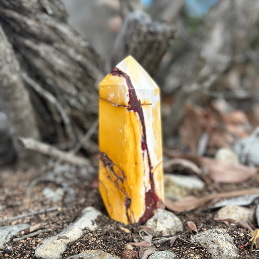A yellow and red mookaite jasper crystal tower standing upright on a natural ground with trees in the background.