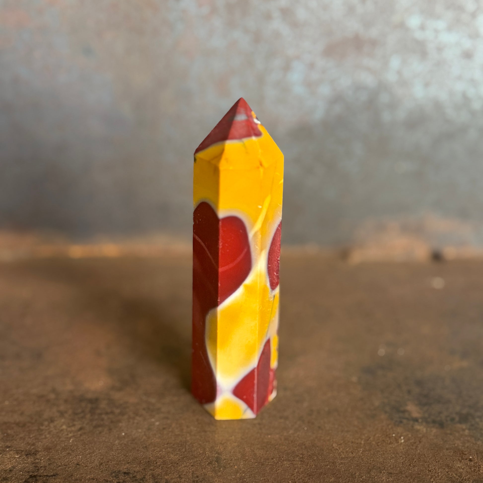 A yellow and red mookaite jasper crystal tower standing upright on a natural ground with trees in the background.
