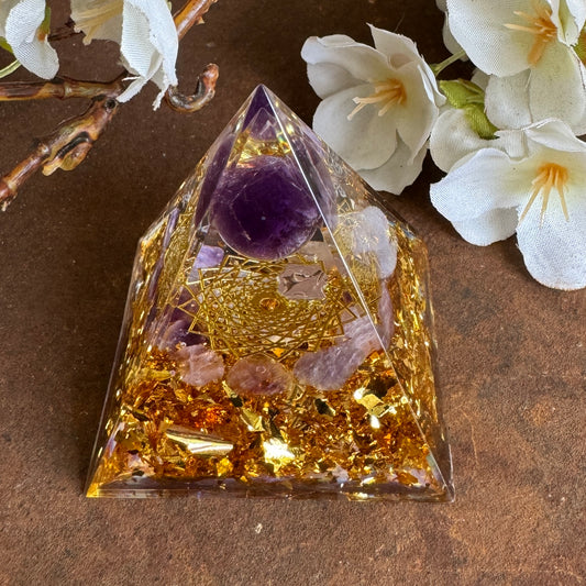 A golden Orgonite Pyramid with Amethyst pieces and a Flower of Life design, placed on a brown surface with white flowers around it.