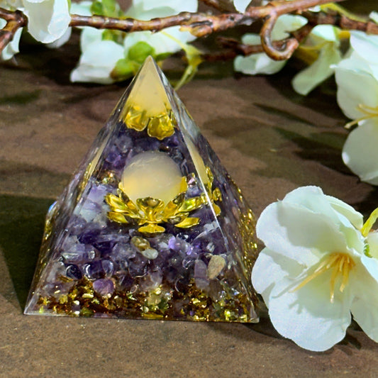 An Orgonite Pyramid made of Amethyst with a Metal Lotus Flower and a Rose Quartz Sphere embedded within, placed on a wooden surface with white flowers around it.