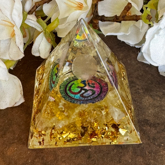 A clear Orgonite pyramid with gold accents and a combination of Citrine, a Clear Quartz Sphere, and an Om Symbol on the top.