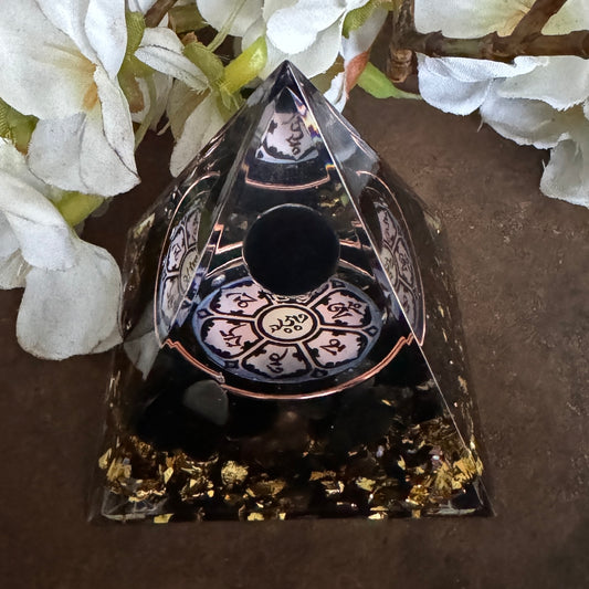 A black Orgonite Pyramid featuring the Om Mani Padme Hum Symbol, placed on a wooden surface surrounded by white flowers and gold confetti.