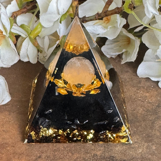 A black Orgonite Pyramid with a Metal Lotus Flower and a Rose Quartz Sphere on top, surrounded by white flowers and green leaves.