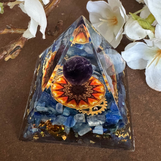 A triangular Orgonite Pyramid with a central Amethyst sphere and Sun feature, surrounded by Kyanite and set against a floral backdrop.