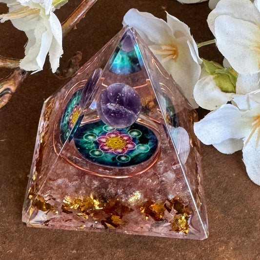 A pink Orgonite Pyramid containing a Rose Quartz, an Amethyst sphere, and a Flower of Life symbol, placed on a wooden surface with white flowers around it.