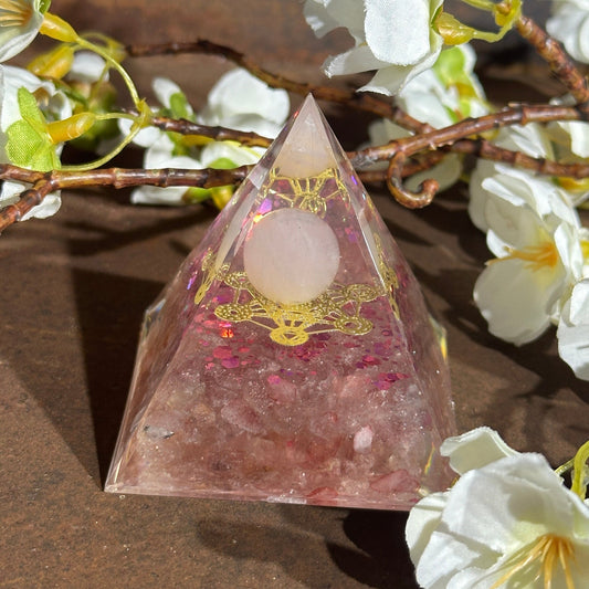 A pink Orgonite pyramid with inclusions of Rose Quartz and Opalite, adorned with a Metal Merkaba Symbol, placed on a wooden surface with white flowers around it.