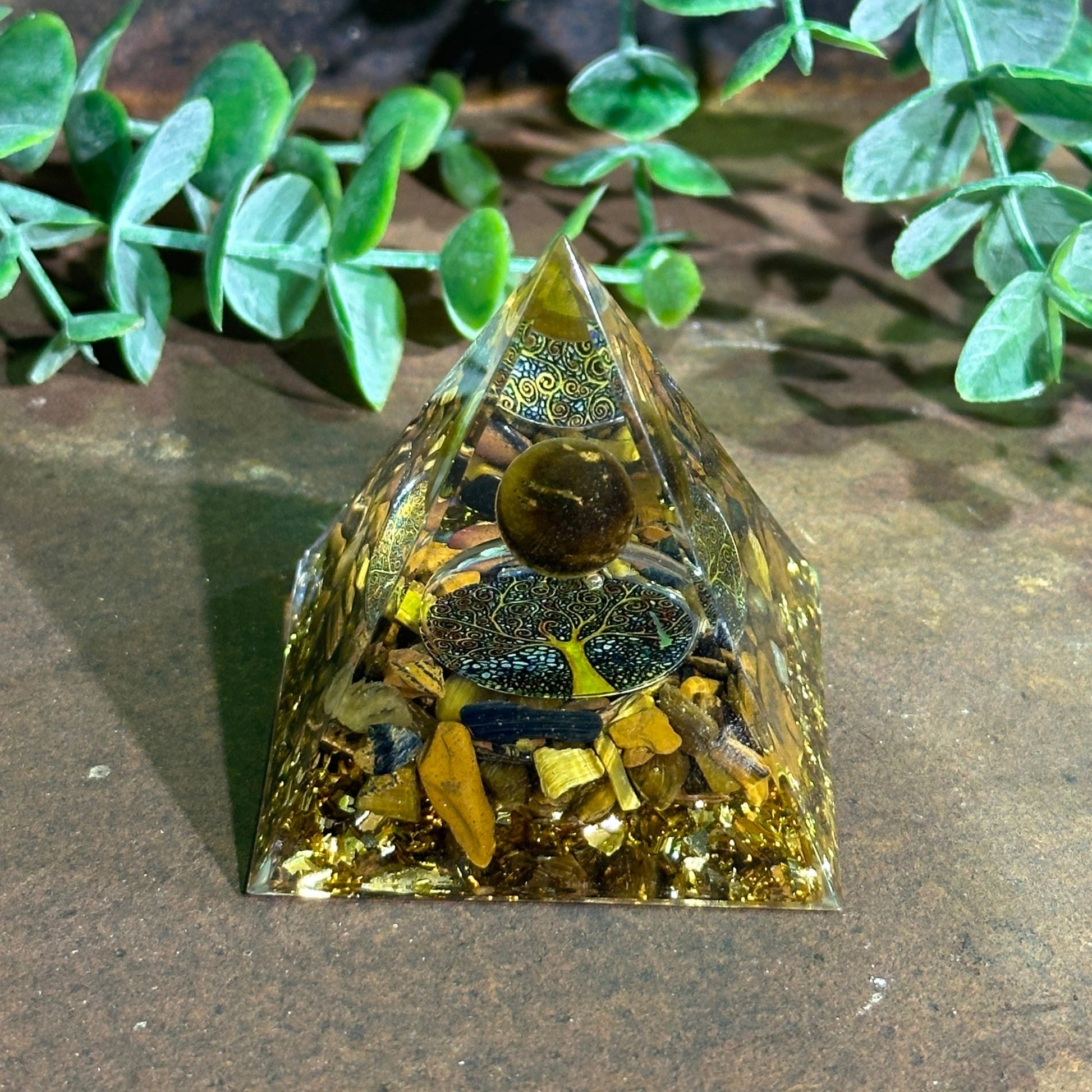 A triangular Organite Pyramid with inclusions of Tiger's Eye gemstone, set against a plant-lined background.