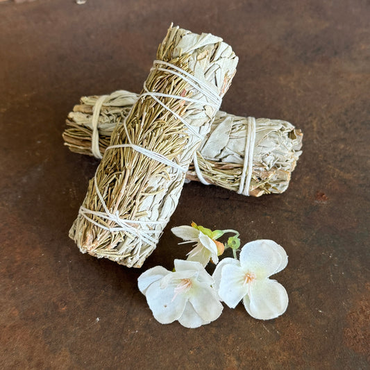 Two bundles of sage tied with twine on a rustic surface with white flowers.