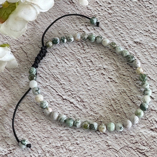 An adjustable bracelet made of Tree Agate beads, displayed on a wooden-like surface with white flowers in the background.